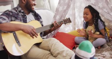 Mutlu Afro-Amerikan baba ve kızı battaniye çadırında oturup gitar çalıyor ve şarkı söylüyor. Evde ailece güzel vakit geçiririz..