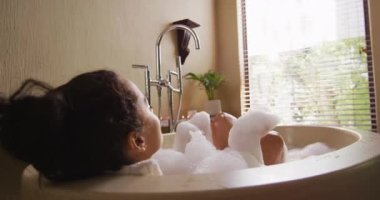 Küvette yatan, köpük üfleyerek kendini şımartan mutlu melez kadın. Güzellik, sağIık ve özgüven, evde boş vakitlerin tadını çıkarmak..