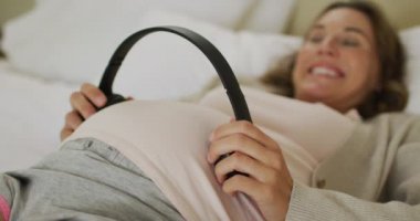 Karnının üstünde kulaklık tutan mutlu, beyaz bir kadının orta kesimi. gebelik, annelik, bebek beklemek, evde zaman geçirmek ve teknoloji konsepti.