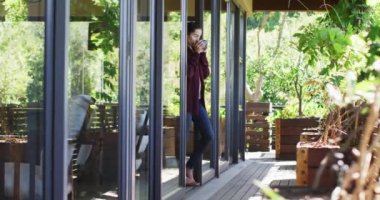 Rahatlamış melez bir kadın kahve içiyor, verandada duruyor, uzaktan bakıyor. ev hayatı ve kişisel bakım, evde boş zamanların keyfi..