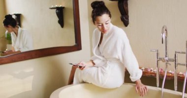 Happy biracial woman sitting in robe on bathtube, using smartphone. beauty, wellness and self care, enjoying leisure time at home with technology.