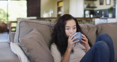 Rahat, melez bir kadın kahve içiyor, kanepede oturuyor. ev hayatı ve kişisel bakım, evde boş zamanların keyfi..