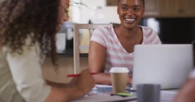 Mutfakta elinde kahveyle oturup laptopla gülen iki farklı kadın arkadaş. Teknoloji ve iletişim, evden esnek çalışma.