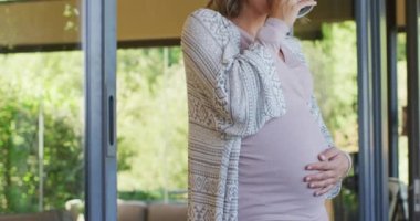 Kafkasyalı hamile bir kadın bir fincan çayla pencerede duruyor, karnına dokunuyor. Hamilelik, annelik, bebek beklemek ve evde zaman geçirmek..