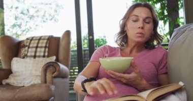 Kafkasyalı hamile kadın kanepede uzanıyor, yemek yiyor ve kitap okuyor. Hamilelik, hamile kalmak ve evde vakit geçirmek..