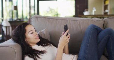 Çift ırklı mutlu bir kadın kanepeye uzanıp akıllı telefon kullanıyor. ev hayatı, teknoloji, evde zaman geçirme.