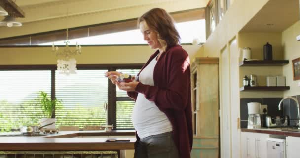 台所に立つ白人妊娠中の女性とアイスクリームを食べる 妊娠中や出産予定日や家で過ごす時間は — ストック動画