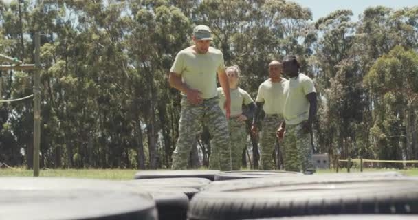 不同的一组士兵看着男教官在阳光下的障碍跑道上跑过轮胎 健康积极的生活方式 在新兵训练营的室外交叉训练 — 图库视频影像