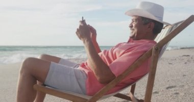 Mutlu İspanyol kıdemli adam gün batımında kumsalda güneşlenirken akıllı telefon kullanıyor. Rahatlayın ve teknoloji ile aktif emeklilik yaşam tarzı.