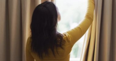 Çift ırklı bir kadının perdeleri açması ve pencereden dışarı bakması. boş zamanlarını evde geçirirler..