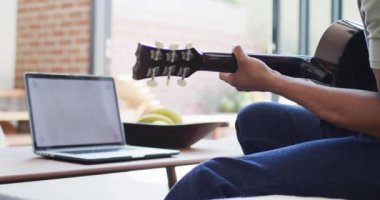 Afrikalı Amerikalı adam evde dizüstü bilgisayar kullanarak gitar çalıp şarkı söylüyor. Teknoloji kullanarak boş zaman geçirmek, evde tek başına dinlenmek..