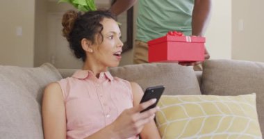 Kanepede oturan çift ırklı bir kadın akıllı telefonuyla erkek partnerinden hediye alıyor. Sevgililer Günü, romantizm ve kutlama.