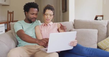 Çift ırklı mutlu çift kanepede oturmuş dizüstü bilgisayarlarıyla konuşuyor. Teknolojiyle evde kaliteli zaman geçirmenin tadını çıkarıyor..