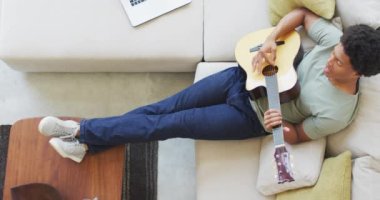 Afrikalı Amerikalı adam evde dizüstü bilgisayar kullanarak gitar çalıp şarkı söylüyor. Teknoloji kullanarak boş zaman geçirmek, evde tek başına dinlenmek..
