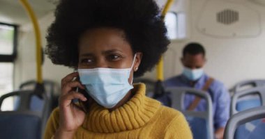 Yüzü maskeli Afro-Amerikalı iş kadını akıllı telefondan konuşuyor ve otobüste oturuyor. Dijital göçebe covid 19 salgını sırasında şehirde dolanıyor..