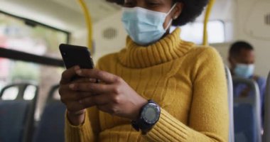 Yüzü maskeli, akıllı telefon kullanan ve otobüste oturan Afrikalı Amerikalı bir iş kadını. Dijital göçebe covid 19 salgını sırasında şehirde dolanıyor..