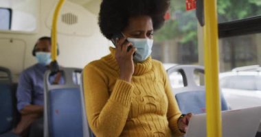 Yüzü maskeli Afro-Amerikalı iş kadını akıllı telefondan konuşuyor ve otobüste oturuyor. Dijital göçebe covid 19 salgını sırasında şehirde dolanıyor..