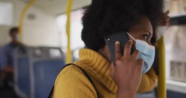 Yüzü maskeli Afro-Amerikalı iş kadını akıllı telefonla konuşuyor ve otobüste duruyor. Dijital göçebe covid 19 salgını sırasında şehirde dolanıyor..