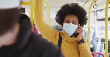 Yüzü maskeli Afro-Amerikalı iş kadını akıllı telefonla konuşuyor ve otobüste duruyor. Dijital göçebe covid 19 salgını sırasında şehirde dolanıyor..