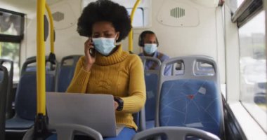 Yüzü maskeli Afro-Amerikalı iş kadını akıllı telefondan konuşuyor ve otobüste oturuyor. Dijital göçebe covid 19 salgını sırasında şehirde dolanıyor..