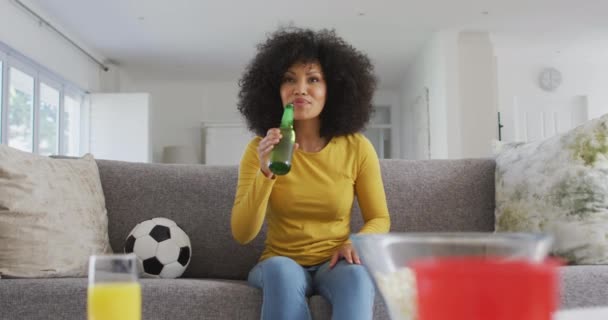 自宅のリビングルームでソファに座って テレビでスポーツを見たり ビールを飲んだり 応援したり 勝利で拳を上げているアフリカ系アメリカ人女性 家で一人で過ごす女 — ストック動画