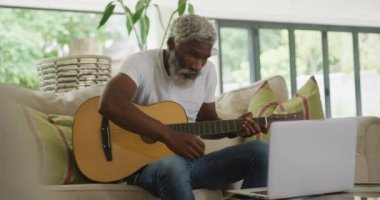 Gitar çalan ve dizüstü bilgisayar kullanan üzgün Afrikalı Amerikalı bir adamın videosu. Emeklilik yaşam tarzı, ev konseptinde dijital olarak oluşturulmuş video ile zaman geçirme.