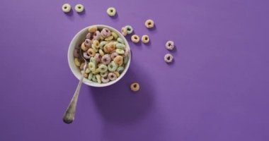 Mor arka planda kâse ve kaşık ile renkli kahvaltı gevrekleri videosu. Kahvaltı ve yemek malzemeleri konsepti.