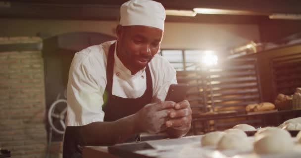スマートフォンを使った幸せなアフリカ系アメリカ人男性パン職人のアニメーション 独立系の中小企業でパン屋で働いています — ストック動画