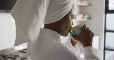 Yüzünde güzellik maskesi olan, mutfakta kahve içen mutlu Afro-Amerikalı kadın. ev hayatı, evde boş zaman geçirmek..