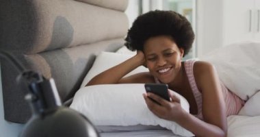 Mutlu Afrikalı Amerikalı kadın yatakta uzanıyor, yatak odasında akıllı telefon kullanıyor. ev hayatı, evde boş zaman geçirmek..