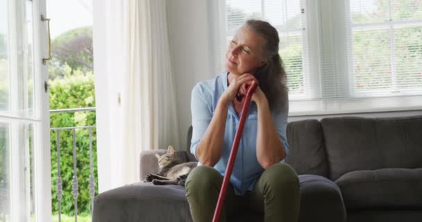 青いシャツを着た上級白人女性がリビングで杖と猫と座っています 家で一人で過ごす — ストック動画