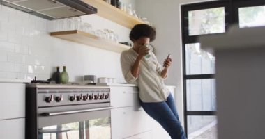 Mutlu Afro-Amerikan kadın kahve içiyor ve mutfakta akıllı telefon kullanıyor. ev hayatı, evde boş zaman geçirmek..