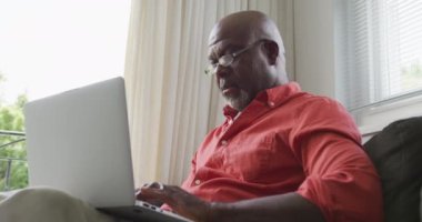 Kıdemli Afro-Amerikan adam oturma odasında dizüstü bilgisayar kullanıyor. Evde yalnız zaman geçirmek..