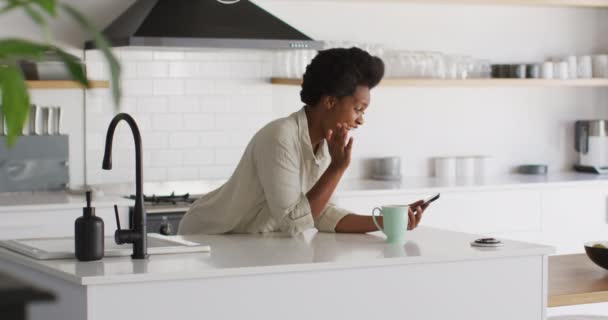 幸せなアフリカ系アメリカ人の女性がコーヒーを飲み キッチンでスマートフォンで話しています 国内での生活や家での自由時間 — ストック動画
