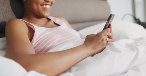 幸せなアフリカ系アメリカ人女性のベッドルームでスマートフォンを使用して ベッドに横たわっていた 国内での生活や家での自由時間 — ストック動画