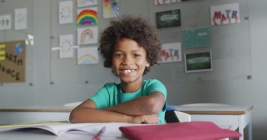Video of happy biracial boy sitting at desk in classroom. primary school education and learning concept.