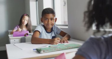 Video of happy biracial boy sitting at desk in classroom. primary school education and learning concept.