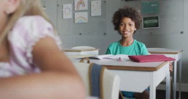 Video of happy biracial boy sitting at desk in classroom. primary school education and learning concept.