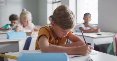Video of focused caucasian boy sitting at desk in classroom. primary school education and learning concept.