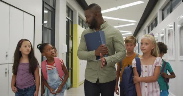 幸せなアフリカ系アメリカ人男性教師と多様な生徒が学校のホールを歩いている映像 小学校教育と教育の専門職 — ストック動画