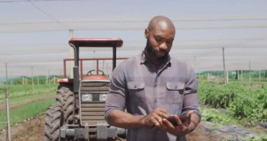 Traktörün önünde elinde tabletle bekleyen Afro-Amerikan bir adamın videosu. Modern organik tarım, tarım sektörü ve teknoloji kavramı.