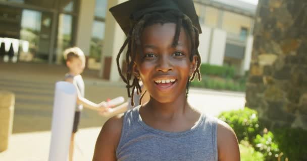 卒業帽子をかぶって学校の前で卒業証書を持っている幸せなアフリカ系アメリカ人の少年のビデオ 小学校教育生態環境ボランティア — ストック動画