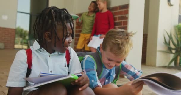 本を持って学校の前で話している幸せな多様な少年たちのビデオ 小学校教育 社会化 — ストック動画