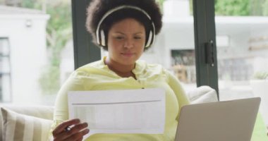 Kulaklıklı, dizüstü bilgisayarlı, koltukta oturan mutlu ve iri yarı Afro-Amerikan kadının videosu. Yaşam tarzı, rahatlık, evde teknolojiyle vakit geçirmek..