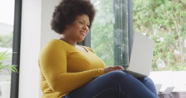 幸せなプラスサイズのアフリカ系アメリカ人女性が床に座ってラップトップを使用してのビデオ ライフスタイルレジャーテクノロジーで家で自由時間を過ごし — ストック動画