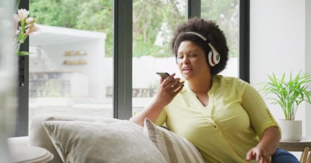 スマートフォンでソファに座ってヘッドフォンで幸せなプラスサイズのアフリカ系アメリカ人女性のビデオ ライフスタイルレジャーテクノロジーで家で自由時間を過ごし — ストック動画