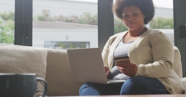 幸せなプラスサイズのアフリカ系アメリカ人女性のビデオクレジットカードを保持し ラップトップを使用しています ライフスタイルレジャーテクノロジーで家で自由時間を過ごし — ストック動画