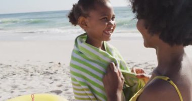 Gülümseyen Afro-Amerikan annesi güneşli plajda kızına havlu atıyor. sağlıklı, aktif aile plaj tatili.