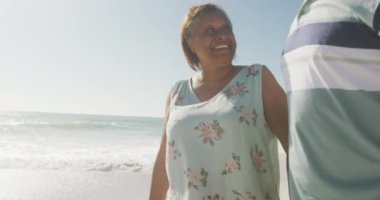 Gülümseyen kıdemli Afrikalı Amerikalı çift güneşli sahilde sarılıp yürüyorlar. sağlıklı, aktif emeklilik plaj tatili.