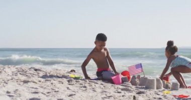 Gülümseyen Afro-Amerikan kardeşler güneşli sahilde Amerikan bayrağıyla kumdan kale inşa ediyorlar. sağlıklı, aktif aile plaj tatili.